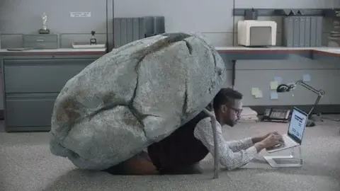 Man in an office cubicle on the floor types on laptop, "sees" the camera observing him, pops out a stick from propping up the large fake "rock", which proceeds to conceal the point of entry into the floor that he might "hide". In other words, he's hiding under a rock.